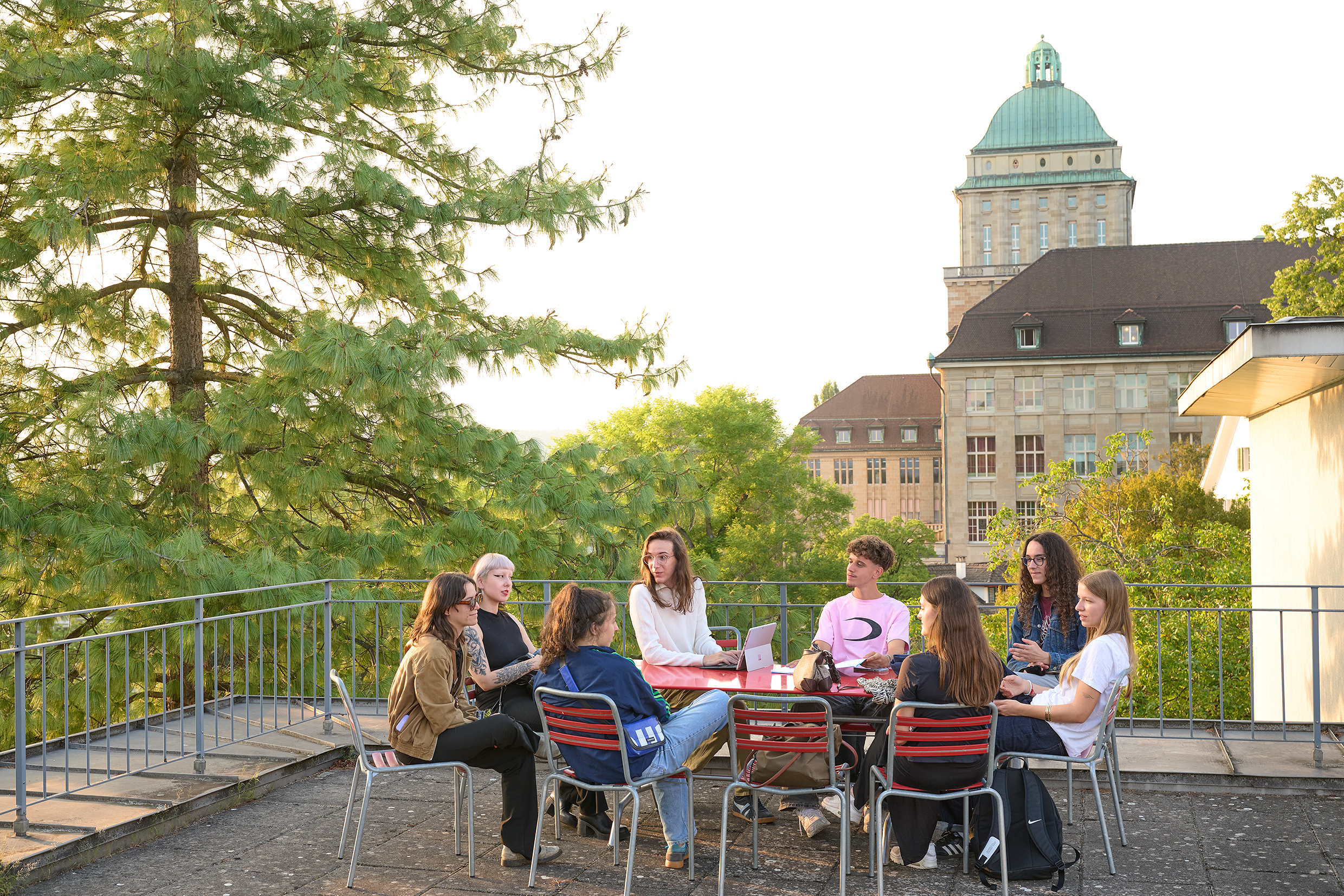 Das Bild zeigt ein einen Eingang des Hauptgebäudes der Universität, eine Gruppe von Studierenden stehen und sitzen auf der Freitreppe und diskutieren.