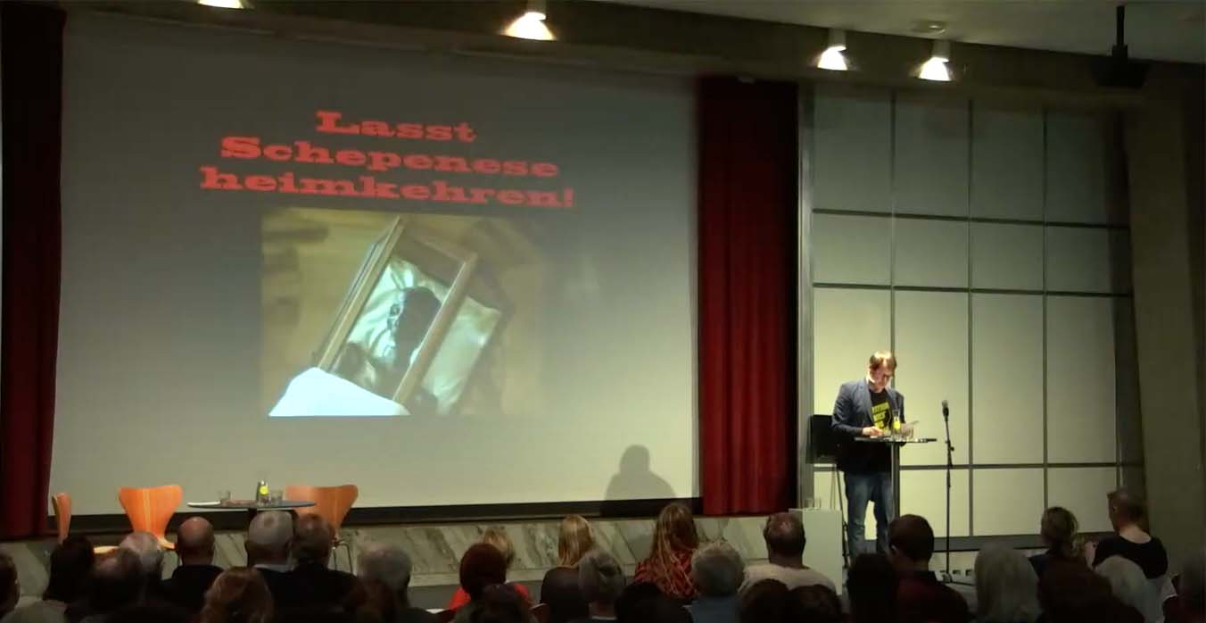 Milo Rau steht rechts auf der Bühne im Vortragssaal des Kunsthauses, hinter ihm ist ein Bild des Sargs der Mumie in der Stiftsbibliothek St. Gallen abgebildet. Darüber steht in roten Buchstaben: Lasst Schepenese heimkehren!
