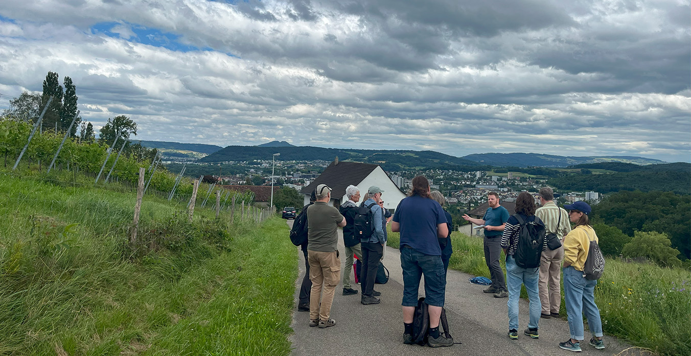 Eine Gruppe von Menschen steht in der Landschaft, grüne Wiesen und ein grauer Himmer.