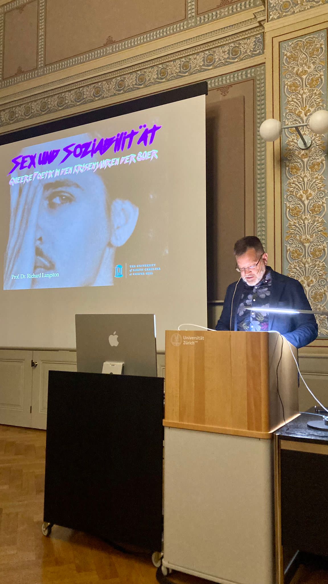 Das Foto zeigt Richard Langston am Rednerpult, im Hintergrund ist ein grosser Screen zu sehen, auf dem ein Mensch sein Gesicht mit einer Hand zur Hälfte abdeckt.