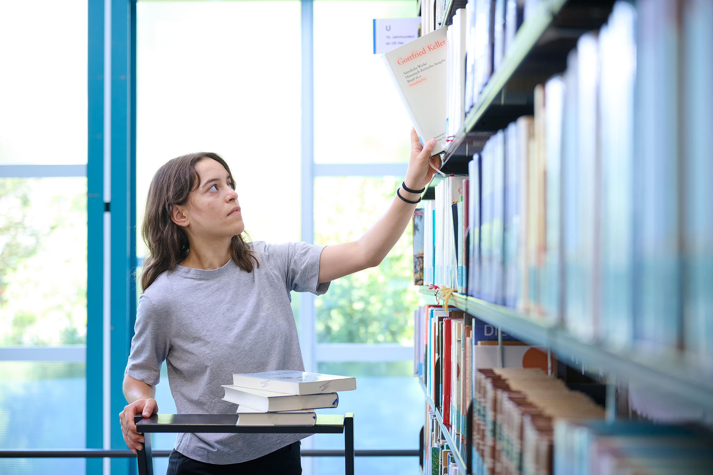 Das Foto zeigt eine Studentin vor einem hohen Bücherregal. Lichtdurchflutet.